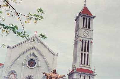The image presents a church building with a prominent clock tower featuring red-tiled roofs and a cross on top. The facade includes an arched window with stained glass. In the foreground, a statue of Jesus with arms outstretched is visible, and branches with green leaves frame the upper left corner.