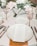 A detailed wedding table setup with white plates and a wedding menu placed on one of them. The table is decorated with elegant floral arrangements and champagne glasses with striped gold stems. Silverware is neatly placed on either side of the plates.