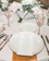 A detailed wedding table setup with white plates and a wedding menu placed on one of them. The table is decorated with elegant floral arrangements and champagne glasses with striped gold stems. Silverware is neatly placed on either side of the plates.
