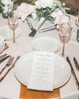 A detailed wedding table setup with white plates and a wedding menu placed on one of them. The table is decorated with elegant floral arrangements and champagne glasses with striped gold stems. Silverware is neatly placed on either side of the plates.
