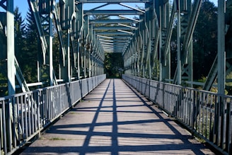A steel bridge with a walkway, featuring a series of evenly spaced beams and railings. Sunlight casts shadows on the path, creating a pattern of lines. Trees and vegetation are visible in the background, adding a natural element to the scene.