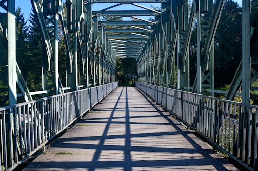 A steel bridge with a walkway, featuring a series of evenly spaced beams and railings. Sunlight casts shadows on the path, creating a pattern of lines. Trees and vegetation are visible in the background, adding a natural element to the scene.