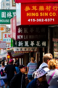 A bustling street scene with people walking along a sidewalk adorned with various brightly colored shop signs, displaying both English and Chinese text. The signs include those for a florist and a company named Hing Sin Co., with vibrant reds and greens being prominent. The street is busy with people of diverse appearances engaging in casual activities.