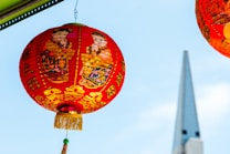 A vibrant red Chinese lantern adorned with colorful illustrations and gold accents, featuring traditional Chinese characters and images of children in festive attire. The lantern is hanging against a clear blue sky, with a pointed architectural structure visible in the background.