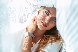 A woman with long blonde hair looks directly at the camera from beneath intricate white lace fabric. Sunlight filters through the lace, casting delicate patterns across her face. She wears a simple white top and a watch with a light-colored strap on her wrist, resting near her neck.