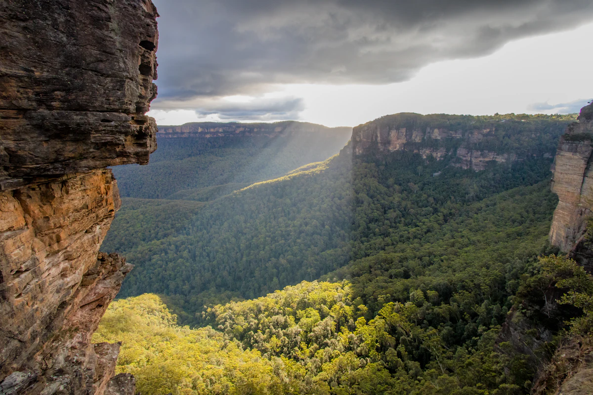 Sandstone cliffs and a forested valley in the Blue Mountains, with sun breaking through storm clouds