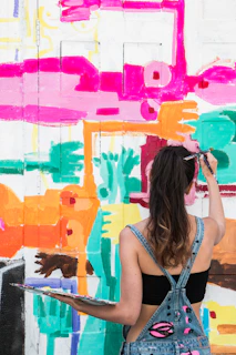 A woman wearing denim overalls and a black top is painting a vibrant mural on a wall. The mural features abstract shapes in bright colors such as pink, orange, turquoise, and yellow. She holds a palette with various paint colors in her left hand while painting with her right hand.