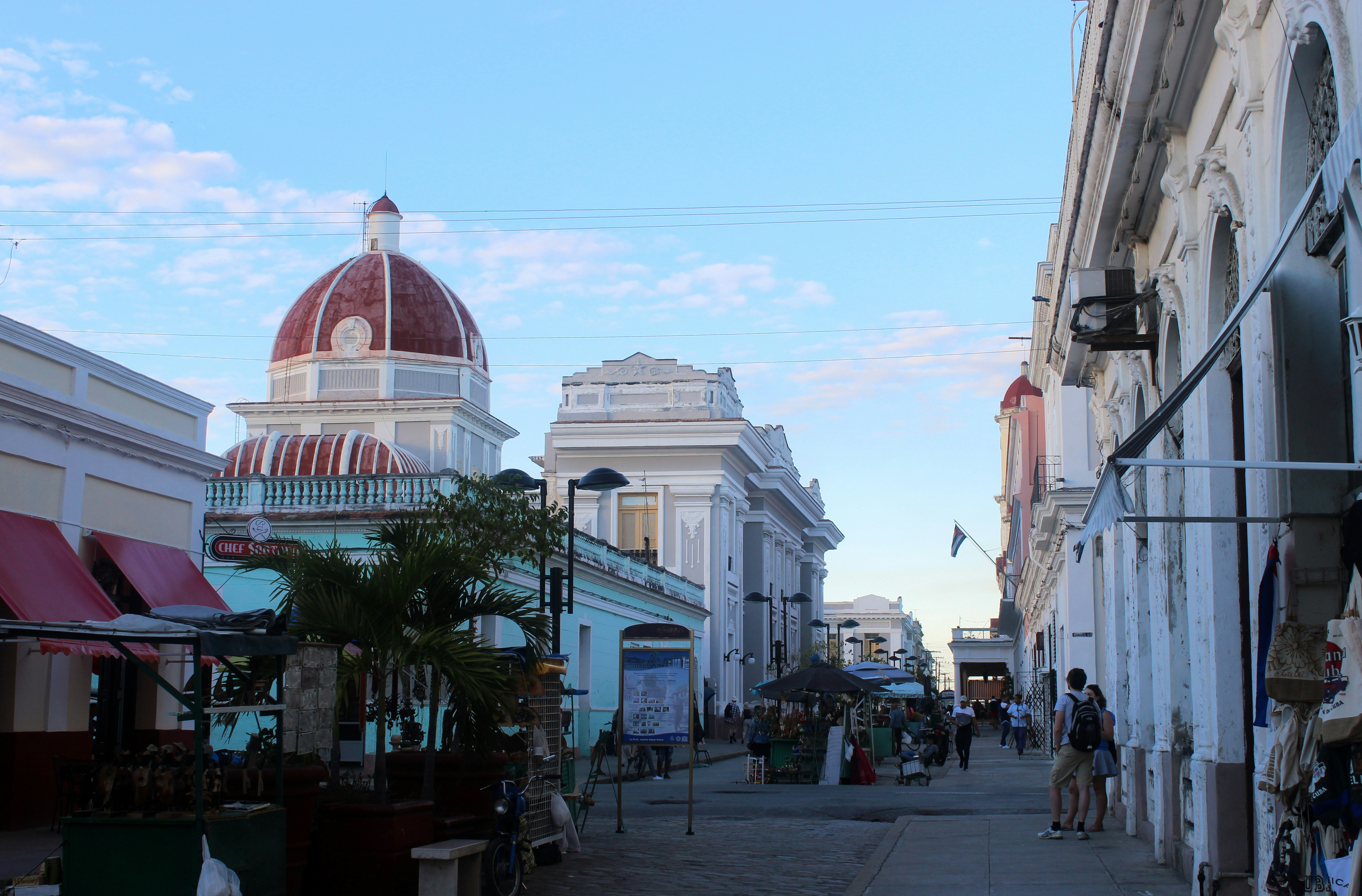 A street scene features colonial-style architecture with a prominent dome topped with a red roof. The area is lively with people walking and exploring various small stalls and shops. Tall buildings line both sides of the street with white and pastel-colored facades, while the sky above is clear and blue with a few scattered clouds.