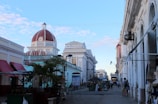 A street scene features colonial-style architecture with a prominent dome topped with a red roof. The area is lively with people walking and exploring various small stalls and shops. Tall buildings line both sides of the street with white and pastel-colored facades, while the sky above is clear and blue with a few scattered clouds.