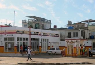 A small, urban gas station with a few people walking nearby. The area has a slightly worn appearance, with old cars parked in front and signage for the gas station, 'Bahia', visible. The building architecture is simple and functional.
