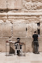 Two individuals are engaged in prayer at a historic stone wall. One is seated on a white chair with a hat and traditional attire, positioned in front of small tables holding books. The other individual is standing, facing the wall, appearing to place a note between the stones.