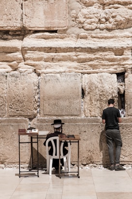 Two individuals are engaged in prayer at a historic stone wall. One is seated on a white chair with a hat and traditional attire, positioned in front of small tables holding books. The other individual is standing, facing the wall, appearing to place a note between the stones.
