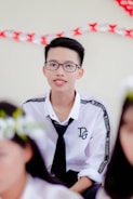A young person with short dark hair and glasses is wearing a white shirt with printed text on the sleeves and a black tie. The background includes decorations with red and white colors.