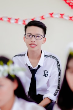 A young person with short dark hair and glasses is wearing a white shirt with printed text on the sleeves and a black tie. The background includes decorations with red and white colors.