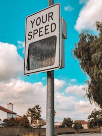 A road sign displays 'YOUR SPEED' at the top and has an electronic display reading 'SLOW DOWN' below. The sign is mounted on a pole against a backdrop of a clear blue sky with scattered clouds. In the background, there are trees and rooftops of suburban houses.