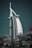 A striking, sail-shaped hotel structure rises prominently against a dark green sky, flanked by lush palm trees at the base. The building features a sleek, modern design with a curved facade and a distinctive cross structure at the top.