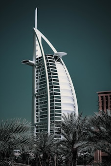 A striking, sail-shaped hotel structure rises prominently against a dark green sky, flanked by lush palm trees at the base. The building features a sleek, modern design with a curved facade and a distinctive cross structure at the top.