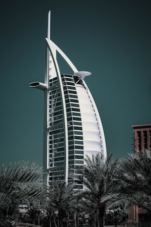 A striking, sail-shaped hotel structure rises prominently against a dark green sky, flanked by lush palm trees at the base. The building features a sleek, modern design with a curved facade and a distinctive cross structure at the top.
