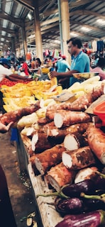 A bustling indoor market scene with stalls displaying various fruits and vegetables. In the foreground, there are large tubers and purple eggplants. Vendors and shoppers are visible in the background, with one seller wearing a blue shirt actively engaged in conversation.