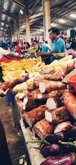 A bustling indoor market scene with stalls displaying various fruits and vegetables. In the foreground, there are large tubers and purple eggplants. Vendors and shoppers are visible in the background, with one seller wearing a blue shirt actively engaged in conversation.