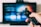 A person holds a remote control in the foreground, with a blurry television screen displaying a user interface in the background. The screen is out of focus, emphasizing the hand and remote control.