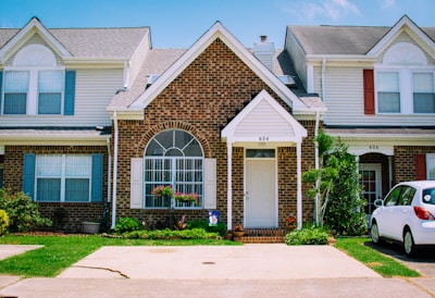 A row of townhouses with brick facades and white trim. Each townhouse has a manicured lawn, potted plants, and decorative elements like arched windows and shutters. A white car is parked in the driveway of one of the houses.