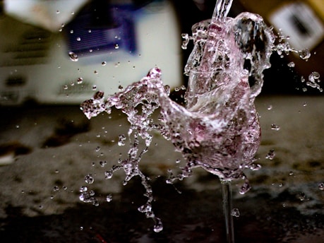 A dynamic splash of liquid captured with remarkable detail, showing droplets flying in multiple directions against a blurred background. The splashes create abstract shapes and convey a sense of movement and fluidity.