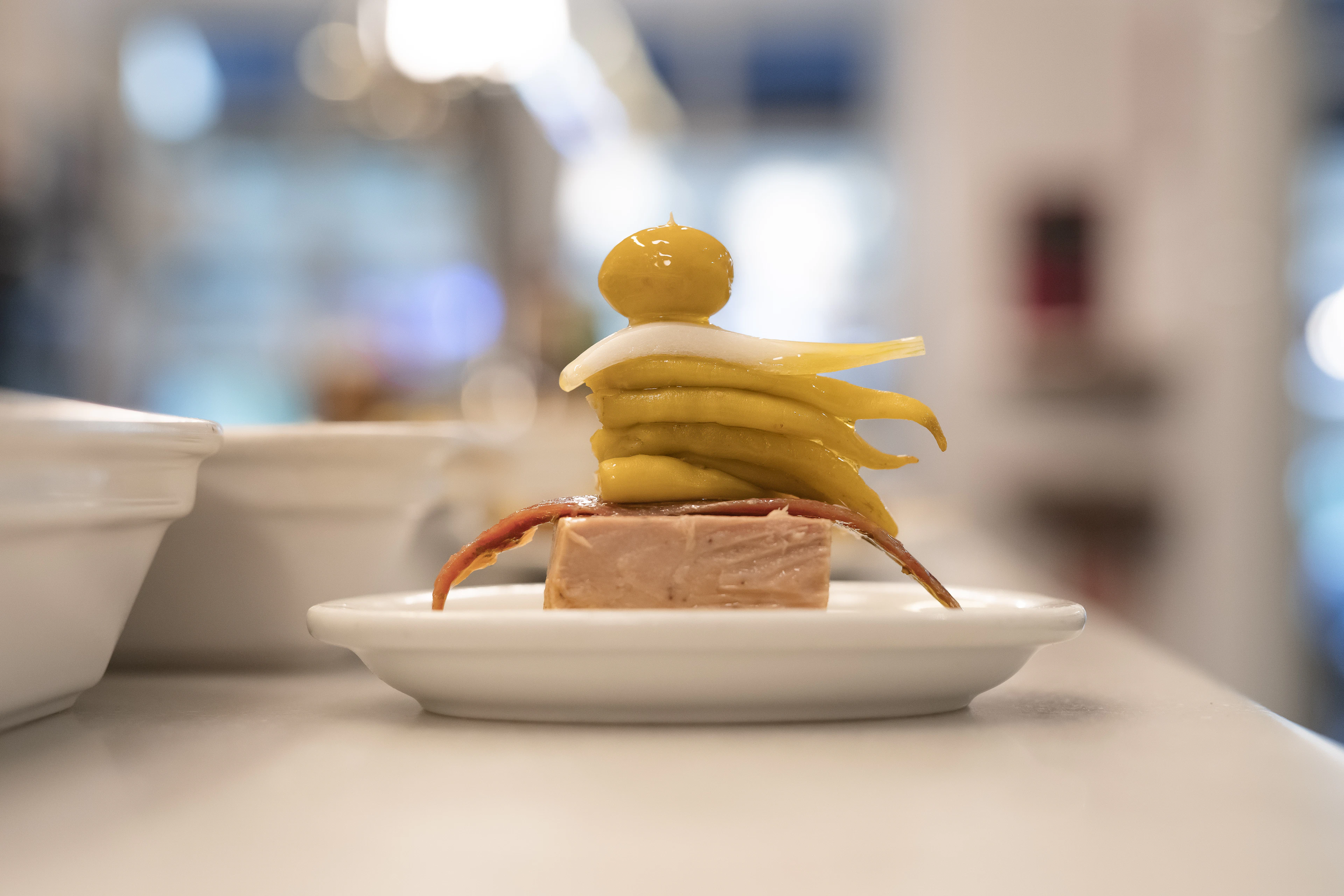 A single pintxo on a small plate on a Basque bar counter, a slab of pate topped with pickled guindilla peppers and an olive