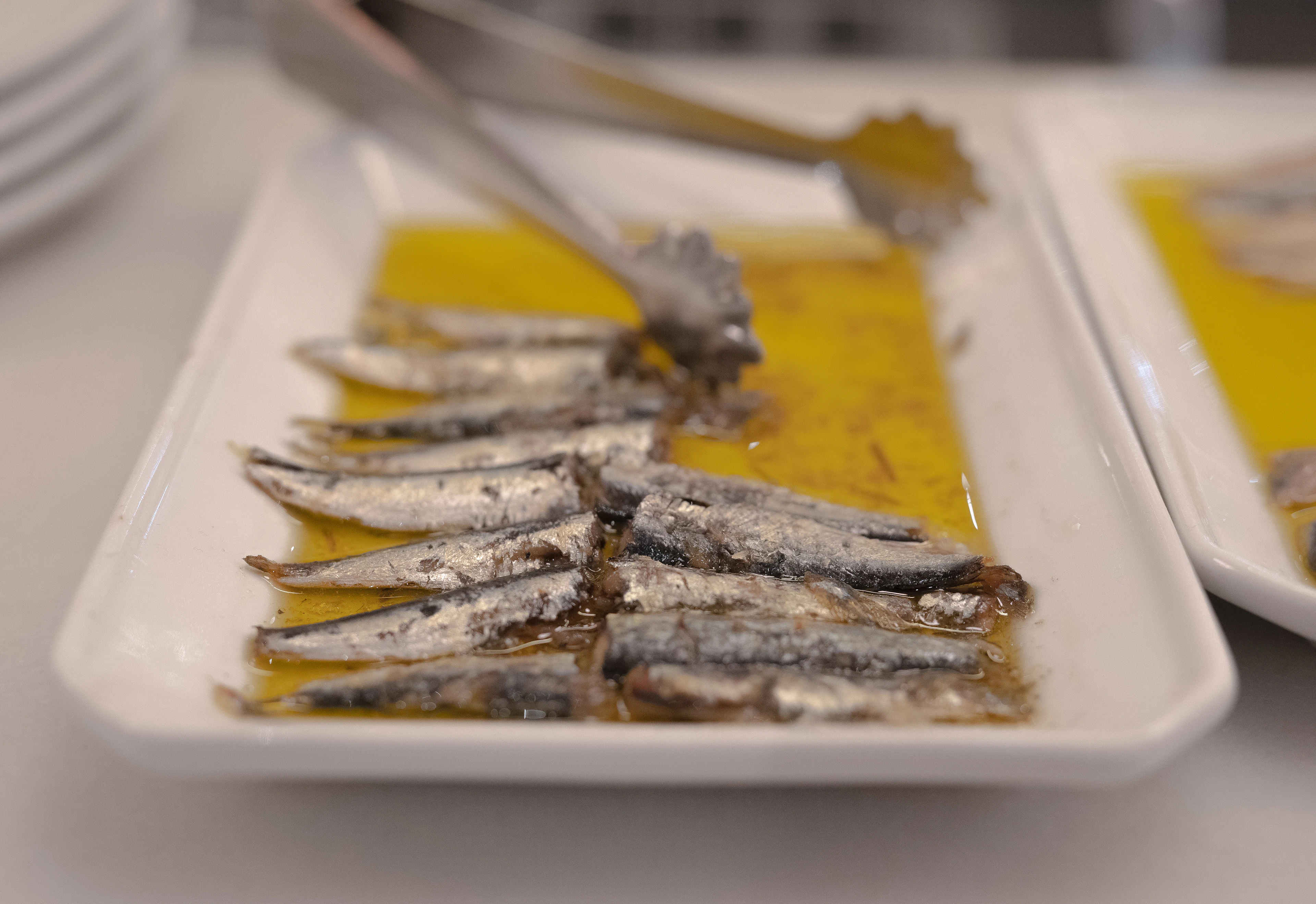 Oil-cured anchovies laid out on white platters with serving tongs, ready to be handed across a Basque pintxo bar counter