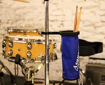 A bright yellow snare drum with chrome detailing on a stand against a textured white brick wall. Beside it, a blue drumstick holder attached to a metal stand holds two wooden drumsticks.