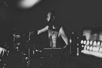 A black-and-white photograph captures a person playing drums in a dimly lit setting. They appear focused, with a moody and artistic atmosphere enveloping the scene. The image highlights the musical environment with emphasis on shadows and contrasts.