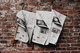Three newspapers are hanging on wooden rods in front of a textured red brick wall. The newspapers feature various articles and advertisements, with some pages partly folded. The brick wall serves as a rustic backdrop, adding a vintage feel to the scene.