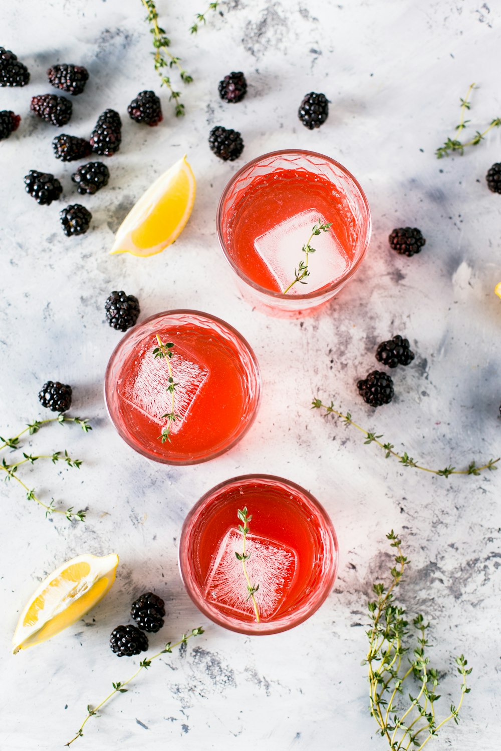 three beverage filled drinking glasses with ice cubes beside sliced lemons and blackberries