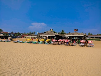 A sandy beach scene with numerous lounge chairs under colorful umbrellas. Several people are relaxing near a beachside café or shack with thatched roofs. The sky is clear with a few clouds, and there are palm trees and other structures in the background.