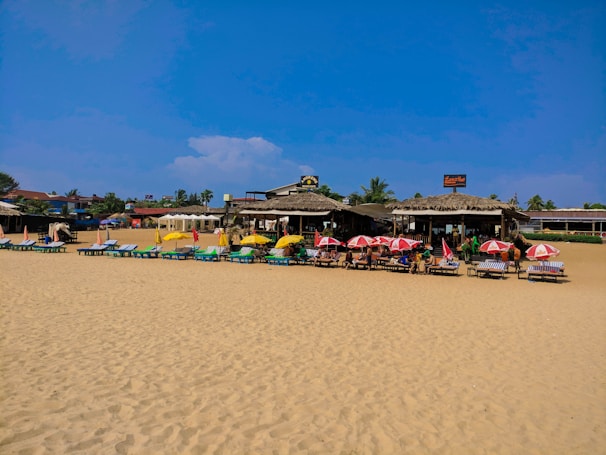 A sandy beach scene with numerous lounge chairs under colorful umbrellas. Several people are relaxing near a beachside café or shack with thatched roofs. The sky is clear with a few clouds, and there are palm trees and other structures in the background.