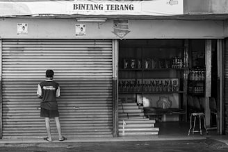 grayscale photo of man standing in front on closed roller shutter