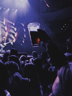 A lively concert scene with a crowd of people enjoying the performance. A person in the foreground holds up a plastic cup filled with beer, silhouetted against the stage lighting. The background shows bright, colorful lights and a musician playing a guitar.
