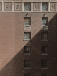A brick building facade with multiple windows arranged in a grid pattern. Ornamental designs are visible at the top near the roofline. A diagonal shadow creates a contrasting effect on the building's surface, adding depth and texture.