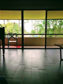 A spacious interior with large, floor-to-ceiling windows opening to a view of a balcony and greenery outside. A single chair and a small table are positioned near the window. The flooring is made of dark tiles and the outdoor area features a wooden deck.