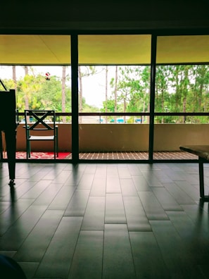 A spacious interior with large, floor-to-ceiling windows opening to a view of a balcony and greenery outside. A single chair and a small table are positioned near the window. The flooring is made of dark tiles and the outdoor area features a wooden deck.
