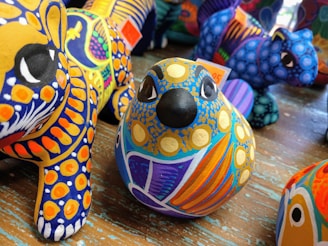 A collection of brightly colored, intricately painted animal figurines crafted in a folk art style. The foreground displays a bird figurine with blue, yellow, and orange patterns, while other figures with similar vibrant designs are visible in the background.