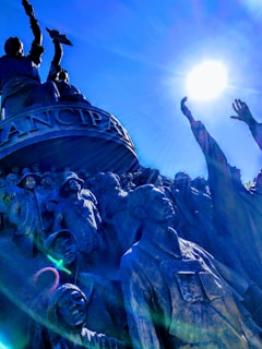 A monument scene featuring a group of statues standing in a powerful and dynamic pose. The figures are depicted in a variety of historical clothing, suggesting a historical or cultural theme. The top figure is raised with an object in hand, reaching towards the bright sun, creating a dramatic interplay of light and shadow.