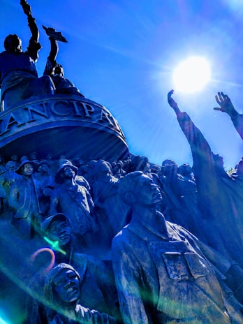A monument scene featuring a group of statues standing in a powerful and dynamic pose. The figures are depicted in a variety of historical clothing, suggesting a historical or cultural theme. The top figure is raised with an object in hand, reaching towards the bright sun, creating a dramatic interplay of light and shadow.
