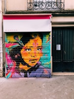 A vibrant and colorful mural depicts a woman's face with elaborate patterns and bright hues. The artwork features bold splashes of color, including pink, orange, blue, and yellow, contrasted against the dark elements of her hair and eyes. The backdrop appears to be a metal roller shutter of a storefront, with a distinct texture visible beneath the paint. Above, there is an awning with a phone number and a wrought-iron railing visible on the building facade.