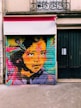 A vibrant and colorful mural depicts a woman's face with elaborate patterns and bright hues. The artwork features bold splashes of color, including pink, orange, blue, and yellow, contrasted against the dark elements of her hair and eyes. The backdrop appears to be a metal roller shutter of a storefront, with a distinct texture visible beneath the paint. Above, there is an awning with a phone number and a wrought-iron railing visible on the building facade.