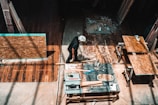 A person is working in a workshop, surrounded by various woodworking tools and materials. There are wooden panels, a measuring square, and a circular saw on the table. The floor is a rich brown wood, and beams of light create patterns on the surface.