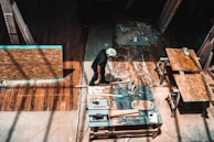 A person is working in a workshop, surrounded by various woodworking tools and materials. There are wooden panels, a measuring square, and a circular saw on the table. The floor is a rich brown wood, and beams of light create patterns on the surface.