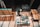 A person is working in a workshop, surrounded by various woodworking tools and materials. There are wooden panels, a measuring square, and a circular saw on the table. The floor is a rich brown wood, and beams of light create patterns on the surface.