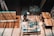 A person is working in a workshop, surrounded by various woodworking tools and materials. There are wooden panels, a measuring square, and a circular saw on the table. The floor is a rich brown wood, and beams of light create patterns on the surface.