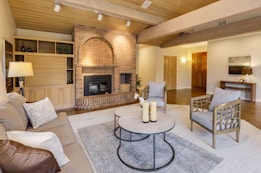 A cozy living room with wooden beams on the ceiling, a brick fireplace, and a mix of modern and rustic furniture. There is a beige sofa with accent pillows, two armchairs with a lattice design, and a round coffee table holding candles. The walls are painted in a light neutral tone and the flooring is wooden. A large rug adds warmth to the space. Shelving units and a small table with a lamp provide additional decor.